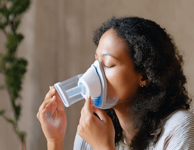 A woman, eyes closed, breathes deeply from a nebulizer, mist enveloping her face, her grip firm on the mask, each inhalation and exhalation a visible battle for air.